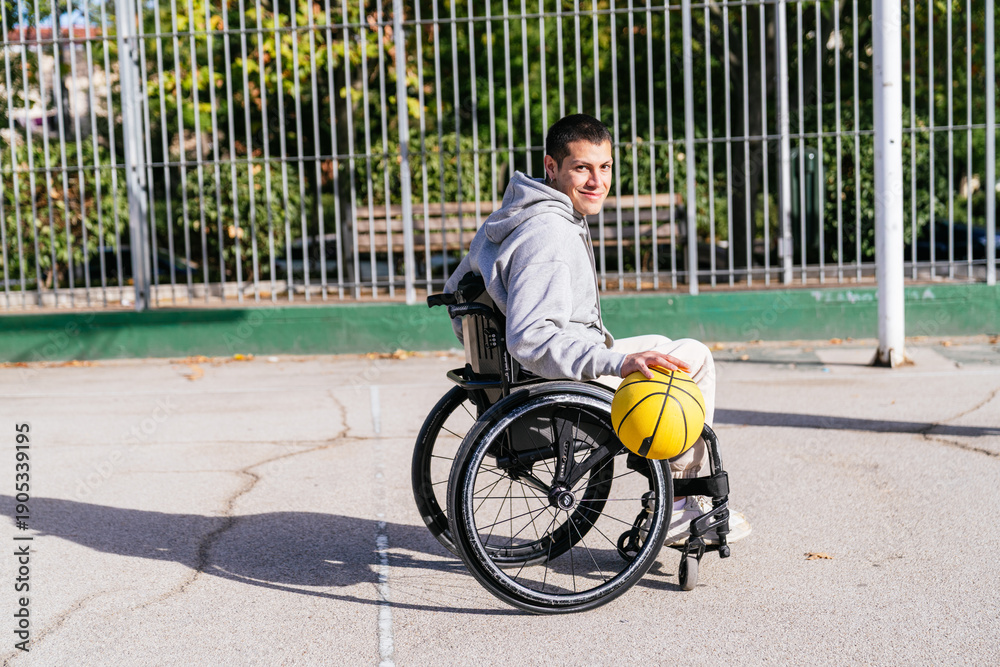 Obraz premium Young man with a prosthetic leg is sitting in a sports wheelchair, holding a basketball on an outdoor court