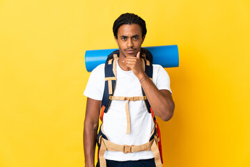 Young mountaineer man with braids with a big backpack isolated on yellow background thinking