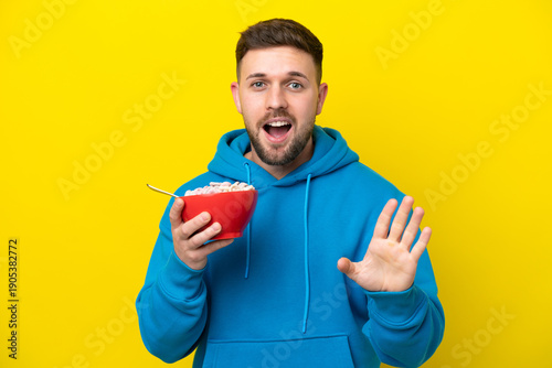 Fotografie Young caucasian man holding a bowl of cereals isolated on yellow background with