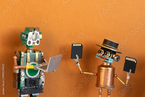 Foto Happy smiling toy robot holding memory cards showing them to another robot with open laptop
