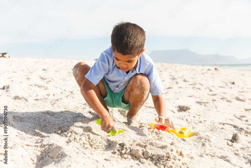Fototapeta premium Full length of boy crouching while playing with sand and toys at beach on sunny day