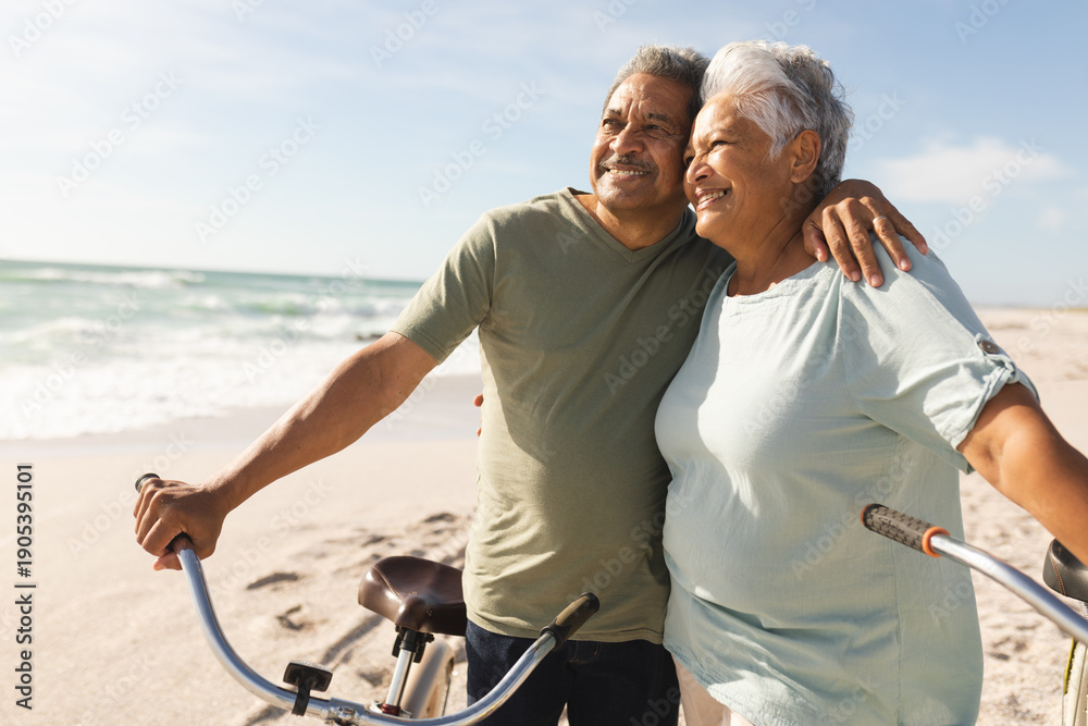 Fototapeta premium Happy senior multiracial couple with bicycles looking away at beach against sky on sunny day