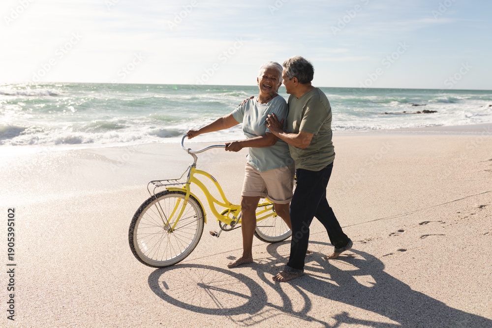 Obraz premium Happy senior multiracial couple talking while wheeling bicycle on shore at beach during sunny day