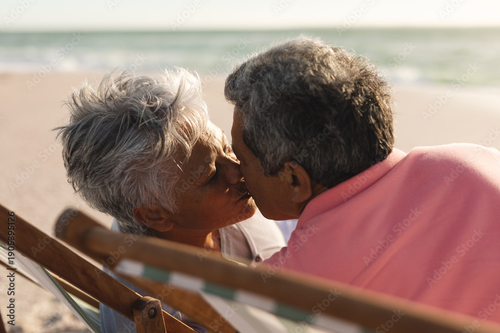 Obraz premium Affectionate senior multiracial couple kissing while sitting on folding chairs at beach