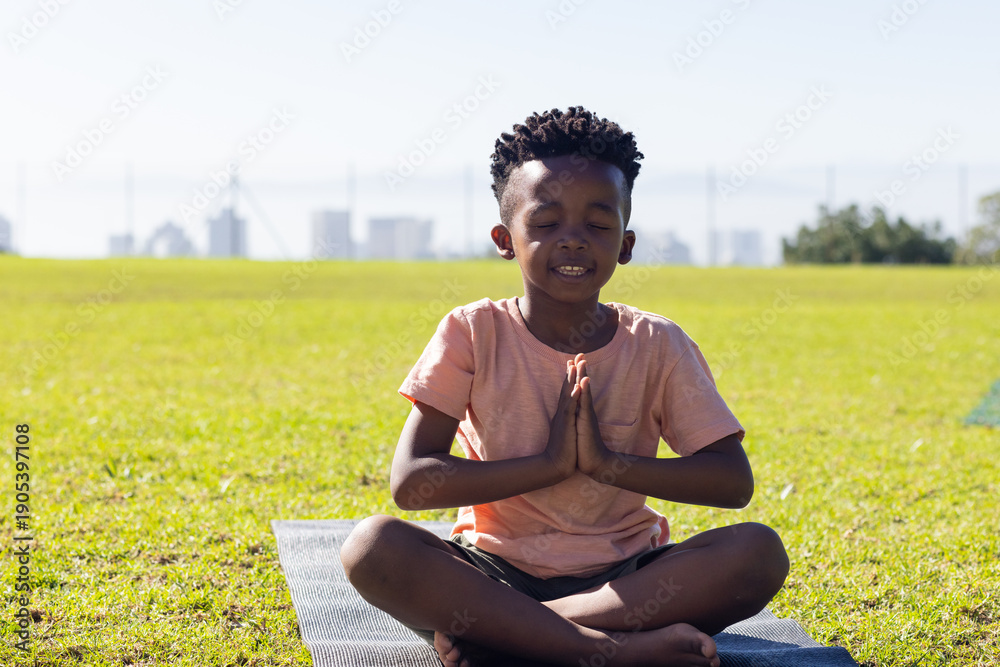 Fototapeta premium Sitting on yoga mat, african american boy meditating outdoors with hands in prayer position