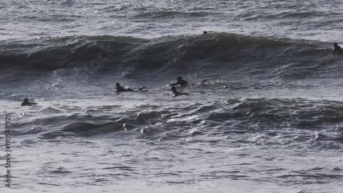 Wide shot of surfers floating in choppy sea as a large wave rolls through. Backlit water surface with spray and swell, outdoor coastal adventure scene.
