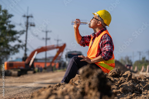 Tired engineer sitting on ground at construction site, feeling hot and thirsty.