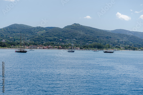 Sailboats on calm sea near coastal town and green hills.