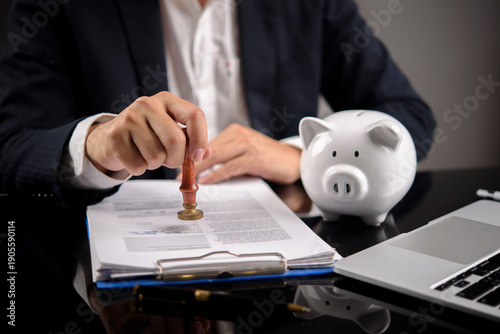 Businessman hand using wooden stamp on contract document near white piggy bank. Concept of loan approval, financial investment agreement, and mortgage savings plan.