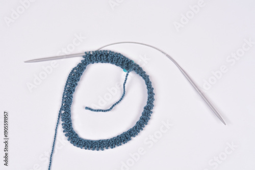 A top down view of a circular knitting project in progress using blue textured yarn and silver circular needles on a clean white surface