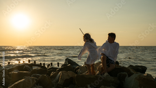Two children, a boy and a girl, sit on coastal rocks at sunset. The children's warm, golden silhouettes rise above the sparkling sea as they play and explore the shore on a carefree summer evening.