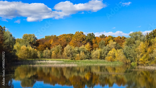 Beautiful autumn landscape in Romania with colorful trees reflecting on a tranquil lake under a blue sky with white clouds.