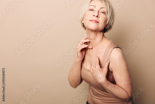 Photography Confident middle aged woman in beige tank top touches her neck and chest, posing against neutral background