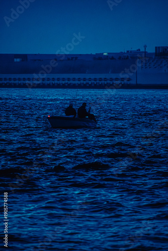 Silhouetted fishermen with fishing rods on a boat drift across dark blue water at dusk. A calm yet dramatic seascape that conveys solitude, friendship, and life at sea.