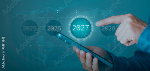 Businessman pointing at 2027 year icon on smartphone. Strategic future planning, business forecasting, long-term goals, and financial roadmap on global world map and graph background.