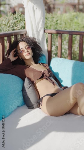 Young woman with curly hair relaxing on a sun lounger by the pool wearing sunglasses
