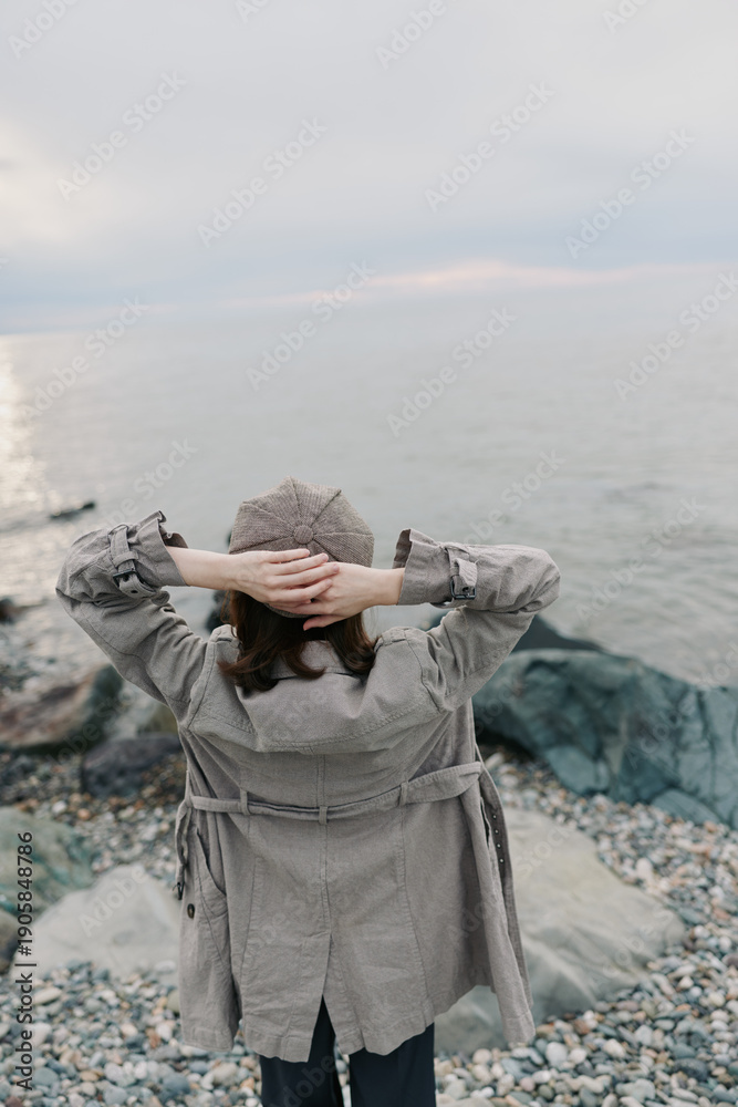 Obraz premium Woman in knitted sweater and hat stands on rocky beach with hands behind head, looking at calm sea under cloudy sky. Concept of relaxation, solitude, autumn, and nature.