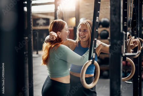 Laughing women standing arm in arm after working out in a gym. One woman has tattoos on her arms, while the other has long red hair.