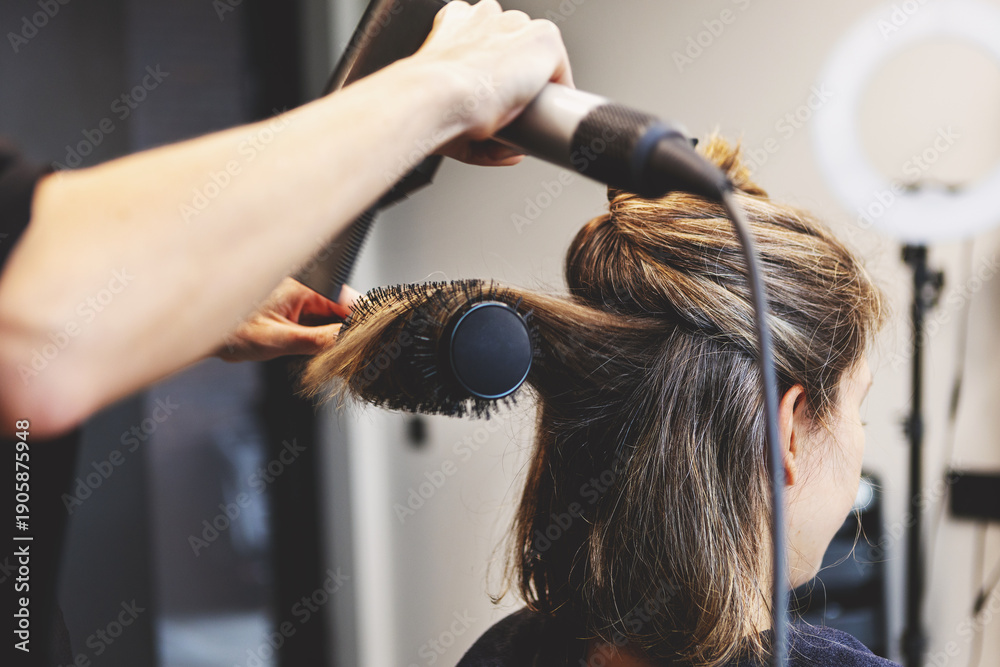 Fototapeta premium Hairdresser blow drying hair of woman with round brush in modern beauty salon, styling sectioned strands. Close up professional hairstyling