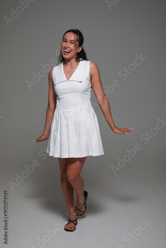 Cheerful brunette young woman with make-up, dressed in a white dress, posing in studio, over grey background. 