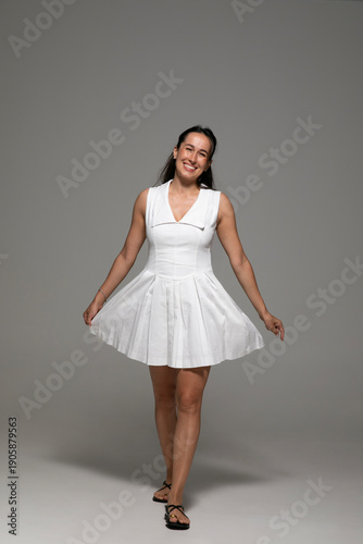 Cheerful brunette young woman, dressed in a white dress, posing in studio, over grey background. 
