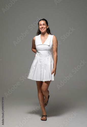 Cheerful brunette smiling young woman with make-up, dressed in a white dress, posing in studio, over grey background. 