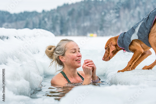 Beautiful blonde hair woman with her dog immersed in the freezing cold water of a lake practicing cold water swimming. Wim Hof Method, cold therapy, breathing techniques, yoga and meditation