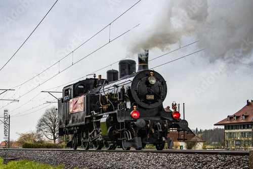 View of a vintage black steam train billowing white smoke against a muted sky, chugging along the tracks near quaint buildings, Lyss, Canton of Bern, Switzerland.