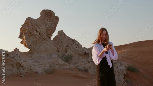 Young caucasian woman in white shirt standing on sand dunes in desert while using smartphone at sunset, representing travel, digital communication and modern lifestyle.