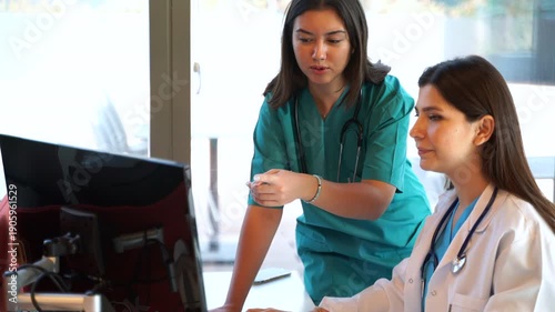 Two female medical professionals, a doctor and a nurse, collaborating and discussing patient data on a computer screen in a clinic office. The doctor takes a break to drink water from a mug.