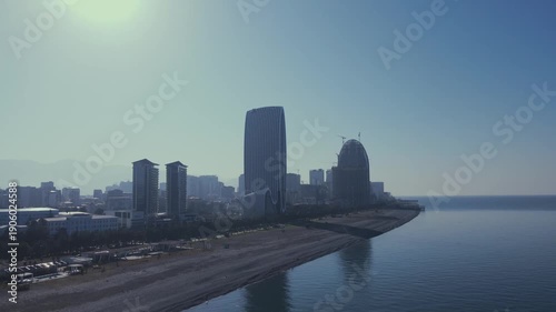 Drone flying backward over Batumi reveals coastline with hotels, residential buildings, calm sea below and mountains on the horizon in soft morning light.