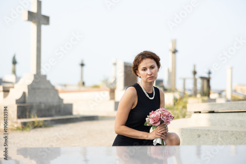 Obraz na plátně Sorrowful young woman in black dress with bouquet of flowers and handkerchief in hands, stands in cemetery