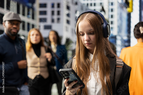 Fotografi Gen z hipster girl walking and scrolling on her mobile phone on the street, wearing big headphones and a backpack
