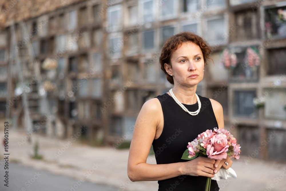 Fototapeta premium Woman in black dress with bouquet of flowers came to cemetery to honor the memory of a deceased loved one