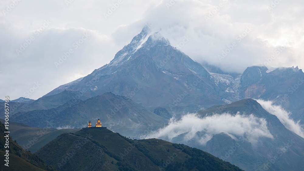 Obraz premium Beautiful summer mountain landscape of the Caucasus, an Orthodox church against backdrop of Mount Kazbek in Georgia, Church illuminated by lights, the mountain peak in clouds