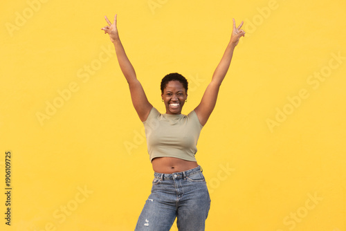 Cheerful young woman with arms raised showing peace sign standing against yellow background. Excited lady in casual clothes showing joyful expression