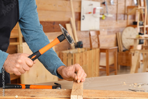 Foto Mature carpenter hammering nail into wooden plank at table in shop, closeup