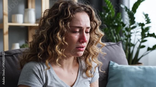 Wallpaper Mural A young woman sitting on a couch in a living room, looking down with a concerned expression, surrounded by plants and a shelf. Torontodigital.ca