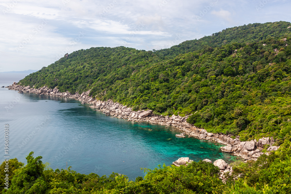 Naklejka premium Lighthouse Bay, Koh Tao, Thailand. Tropical rocky coastline with clear blue water. Lush green forest and scenic seascape. Ocean meets the shore under a cloudy sky.