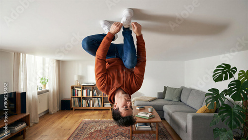 Young man hanging upside down from the ceiling, tying shoelaces in a modern living room for an unconventional lifestyle concept.