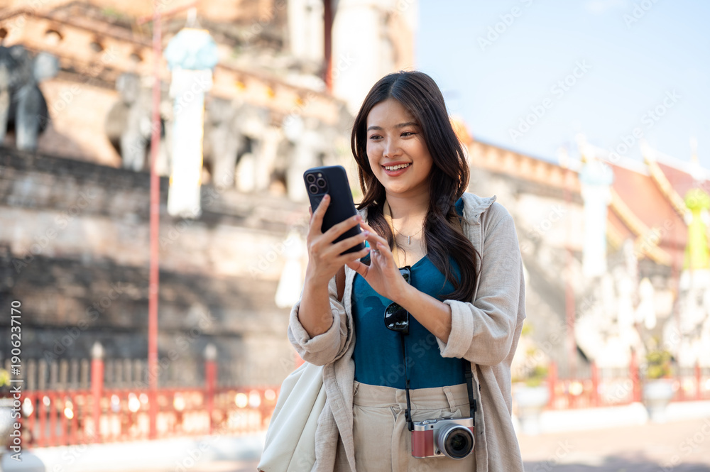 Fototapeta premium Pretty asian woman looking at smartphone while standing aside the ancient pagoda of buddhist temple.