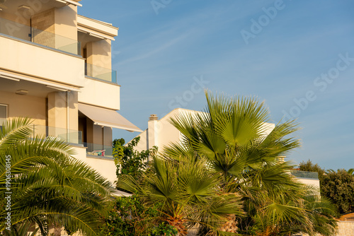 Modern resort hotel building with balconies viewed through green palm trees under blue sky. Concept of summer accommodation, holiday rental, tourism industry, luxury stay and coastal living. 