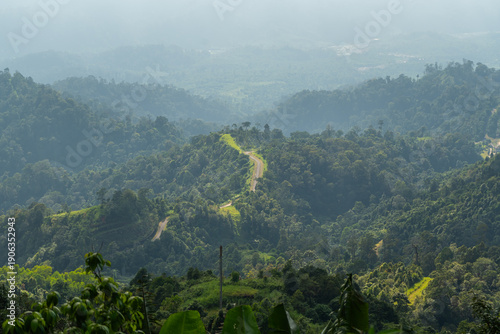 A winding mountain road curves through a lush tropical forest landscape under a hazy sky
