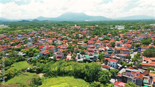 Drone shot over Duong Lam ancient village in Vietnam, showing traditional red-tiled roofs, lush green vegetation, and the majestic Ba Vi mountain range in the background under a bright sky