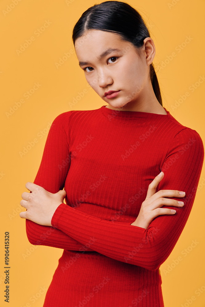 Obraz premium Portrait of young woman with straight black hair in red sweater crossing arms and looking serious on solid yellow background. Asian female model posing with natural expression in studio light.