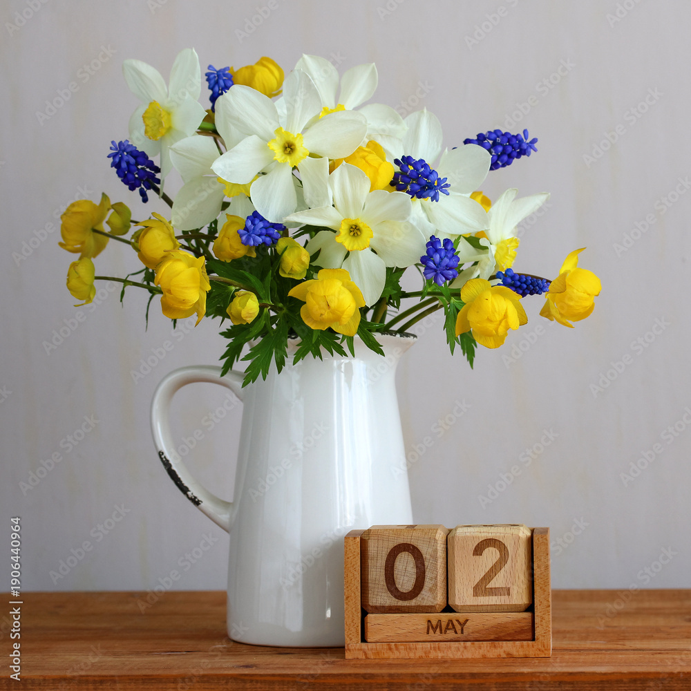 Naklejka premium spring bouquet of daffodils, muscari, and trollius in a white pitcher standing on a wooden surface next to a vintage calendar displaying May 2nd, symbolizing seasonal renewal