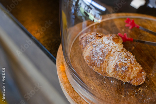 Single croissant resting on round wooden board under dome cover with metal tongs on bakery counter
