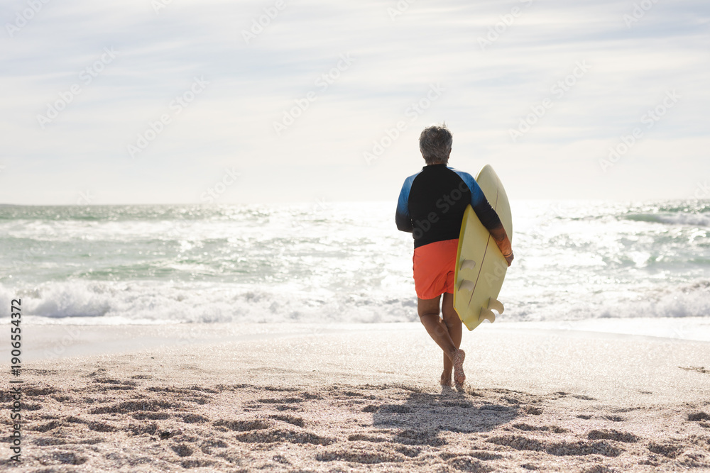 Naklejka premium Full length rear view of senior woman running with surfboard towards sea at sunny beach