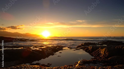 Coastal sunset seascape with rocky shoreline reflecting warm sunlight and serene ocean waves under a vibrant orange sky