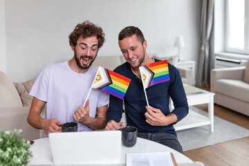 Happy gay men sitting in living room with small pride flags and laptop. LGBT couple laughing while looking at screen. Home lifestyle, diversity and queer representation. Two partners with flags.